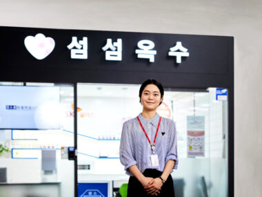 육, 칠, 팔, 구 등 짐칸을 배경으로 빨간색, 파란색, 노란색, 청록색 캐리어가 있고, 여성이 짐을 확인하는 일러스트 그림