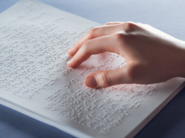 하얀 책 위에 표시된 점자를 손으로 만지고 있는 모습의 사진